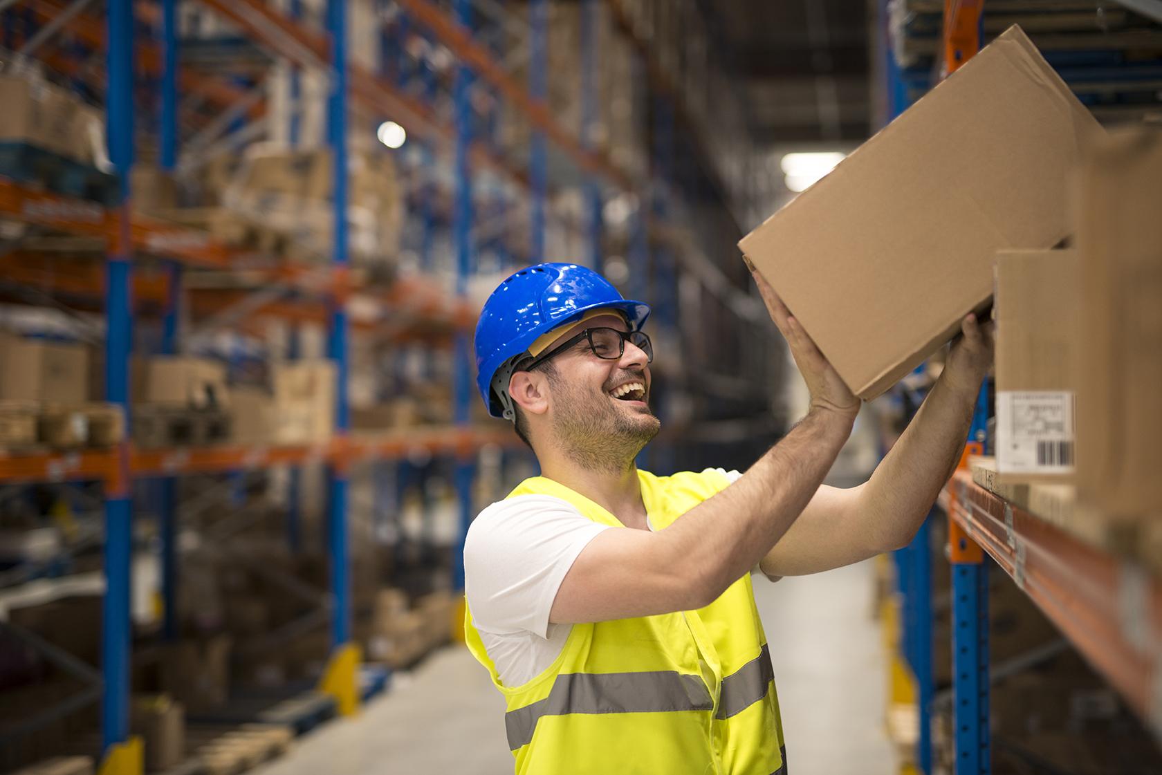 A man in a safety vest and helmet smiling while placing a box on a warehouse shelf.