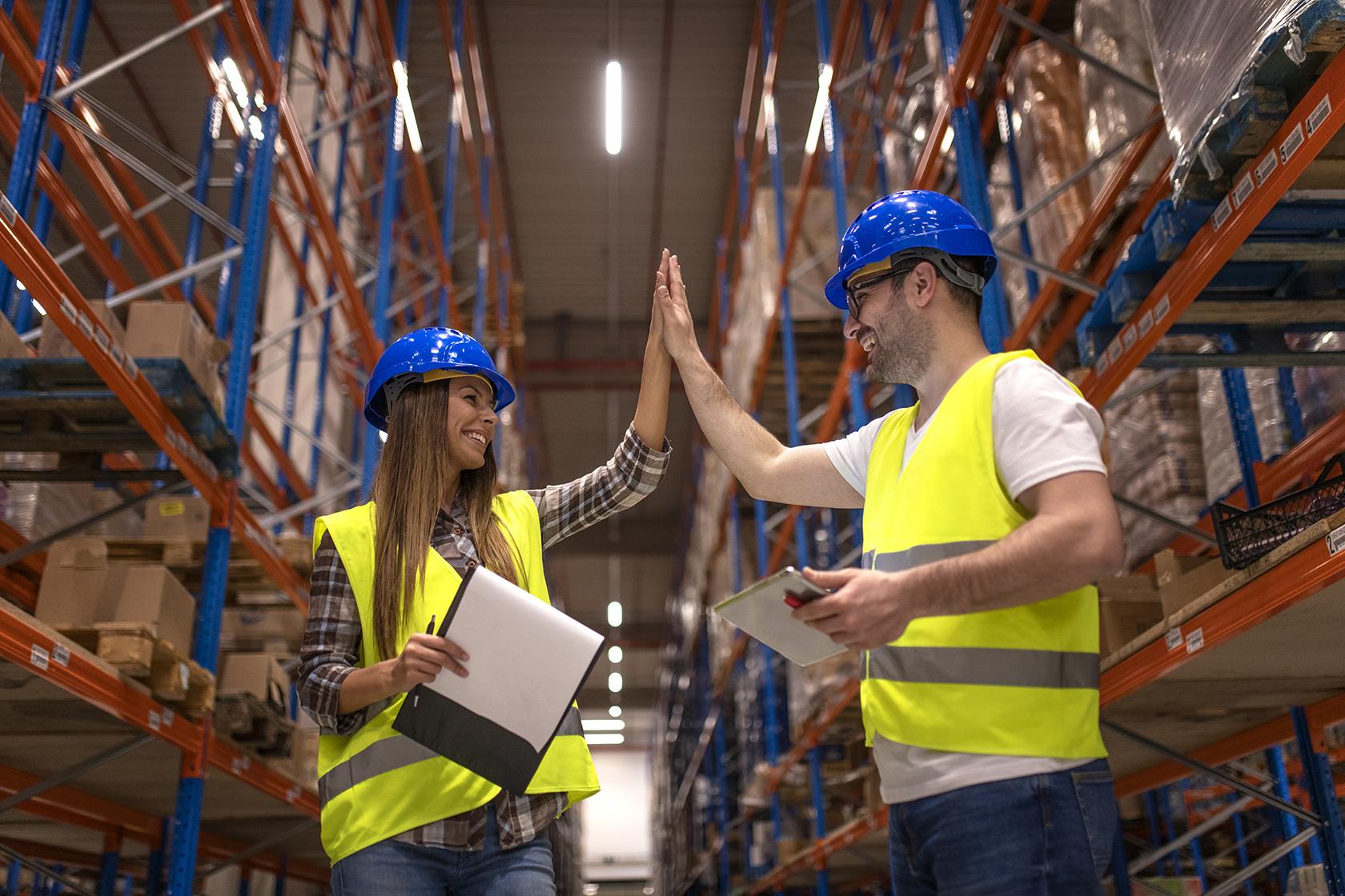Two warehouse workers in safety vests and helmets high-fiving while holding clipboards.
