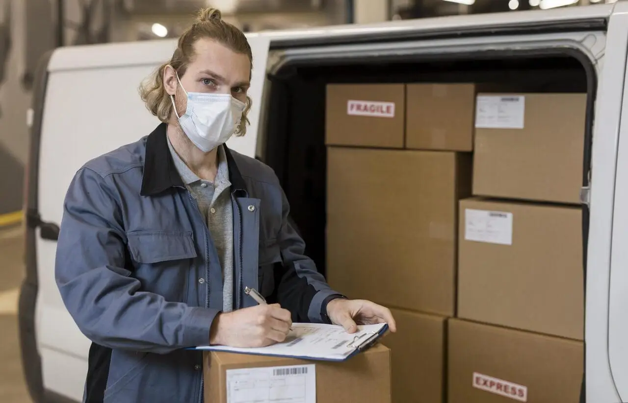 A delivery worker wearing a mask signs paperwork next to a van loaded with boxes.
