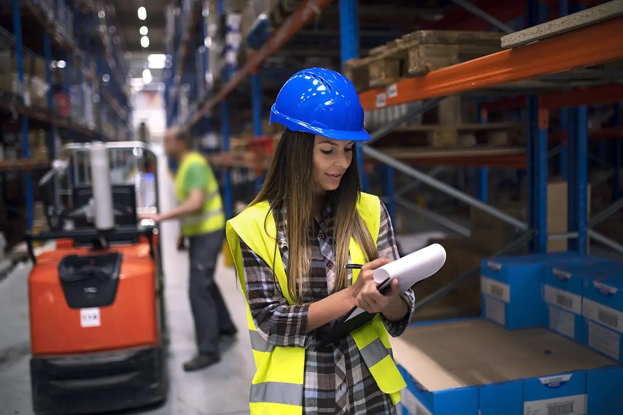 A female worker in a helmet and vest writes on a clipboard in a warehouse.