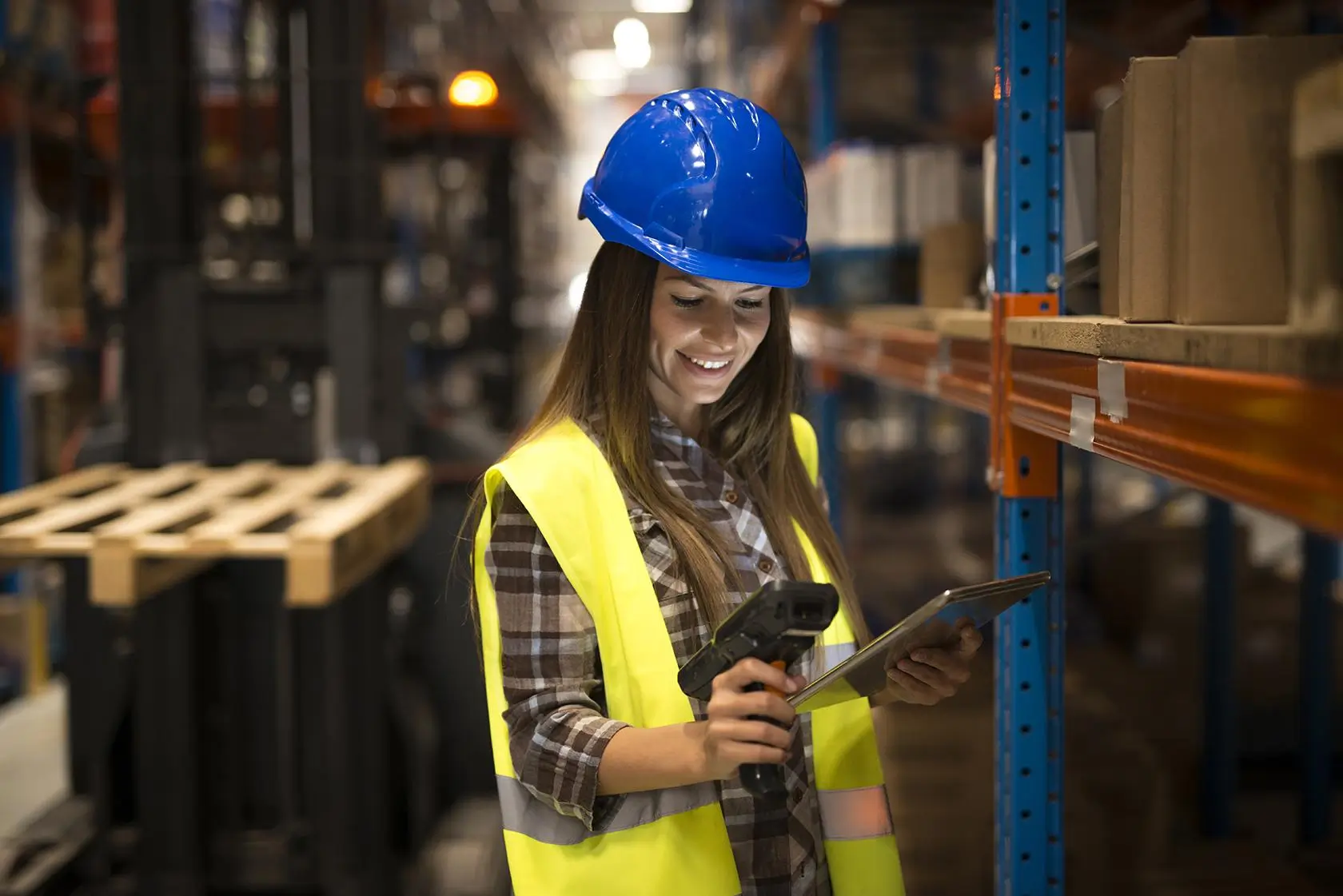 A female warehouse worker in a helmet scans a product while holding a tablet.
