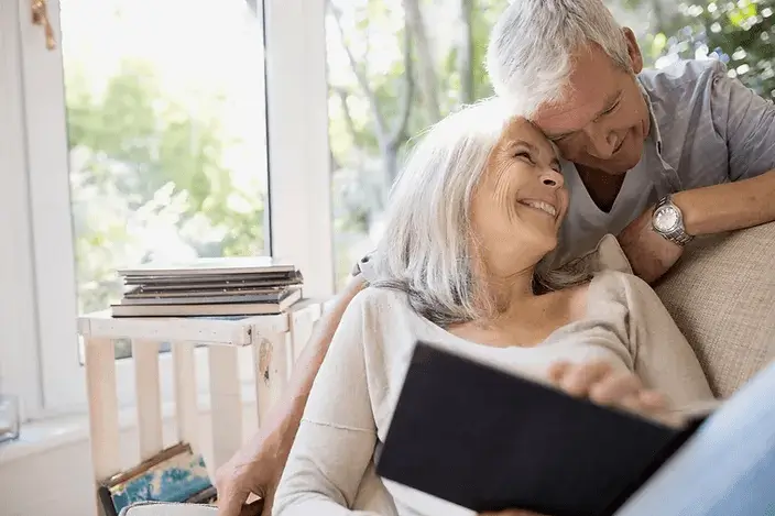 An elderly couple cuddling and smiling while reading a book together.