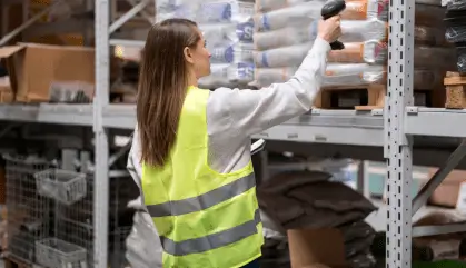 Woman scanning products on a shelf in a warehouse, wearing a yellow safety vest.