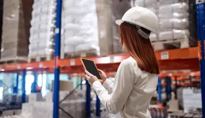Woman in a white helmet using a tablet in a warehouse with tall shelves.