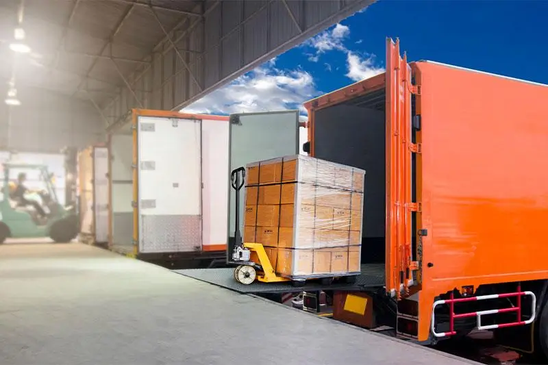 A pallet of boxes is being loaded into an orange truck at a warehouse dock.