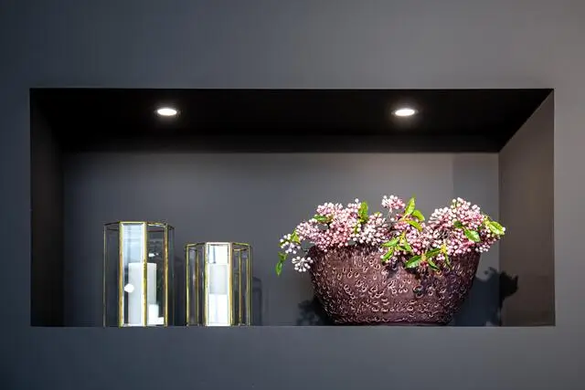 A vase with pink flowers and two glass lanterns are displayed in a recessed shelf with lights.