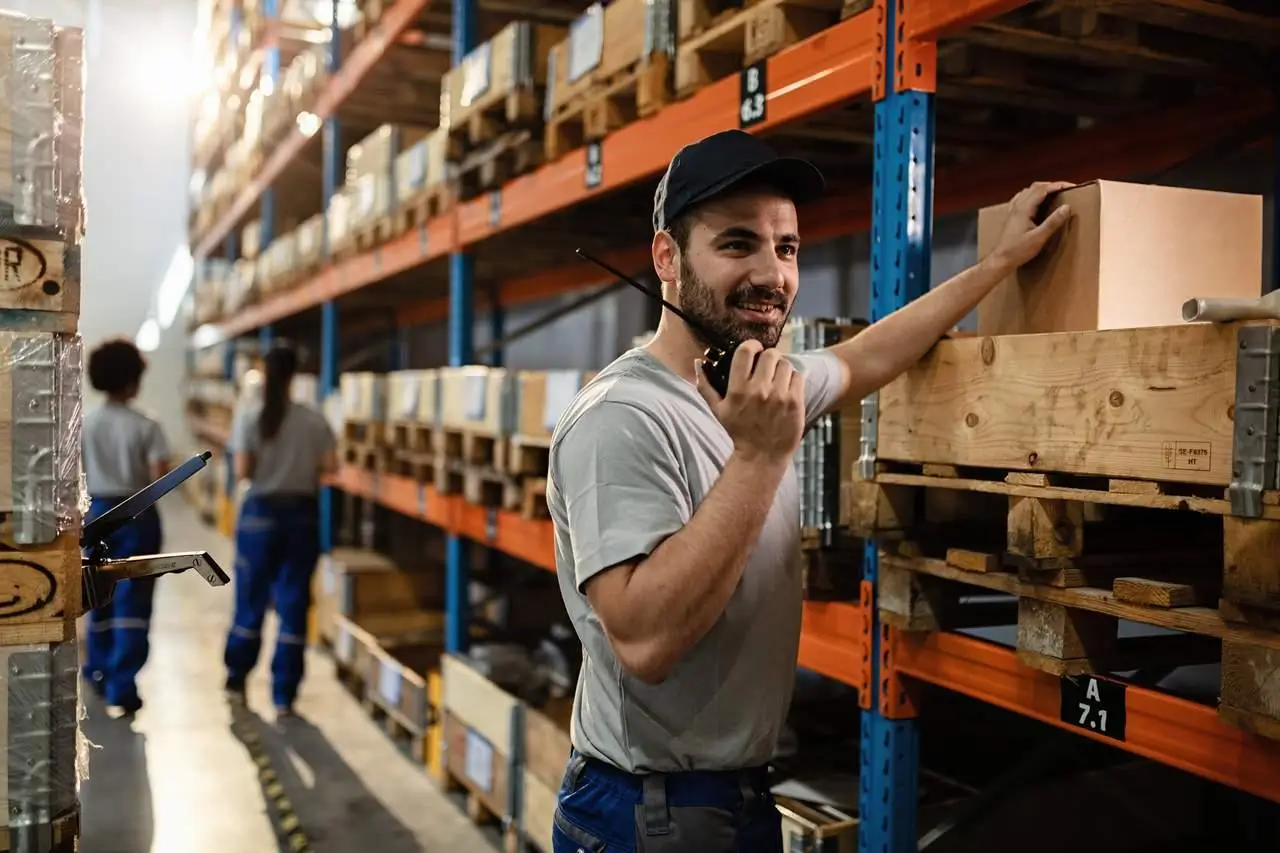 Worker using a walkie-talkie while standing near shelves in a warehouse.