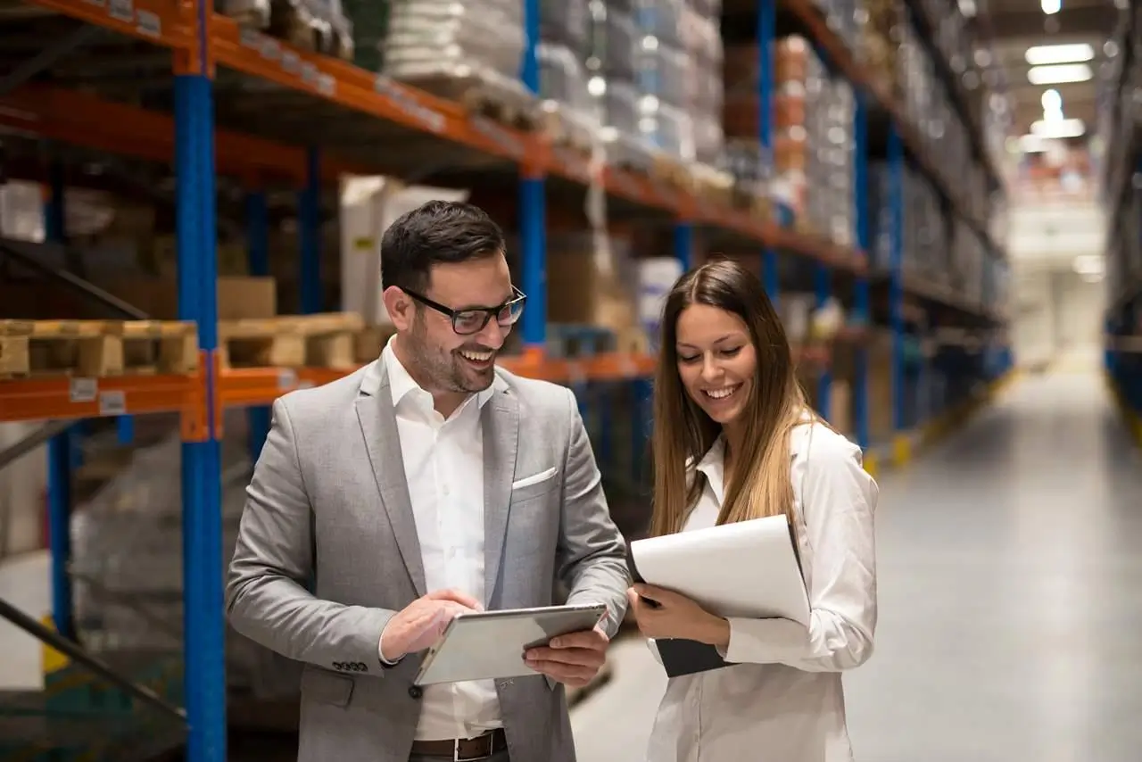 Two people smiling while reviewing documents and a tablet in a warehouse aisle.