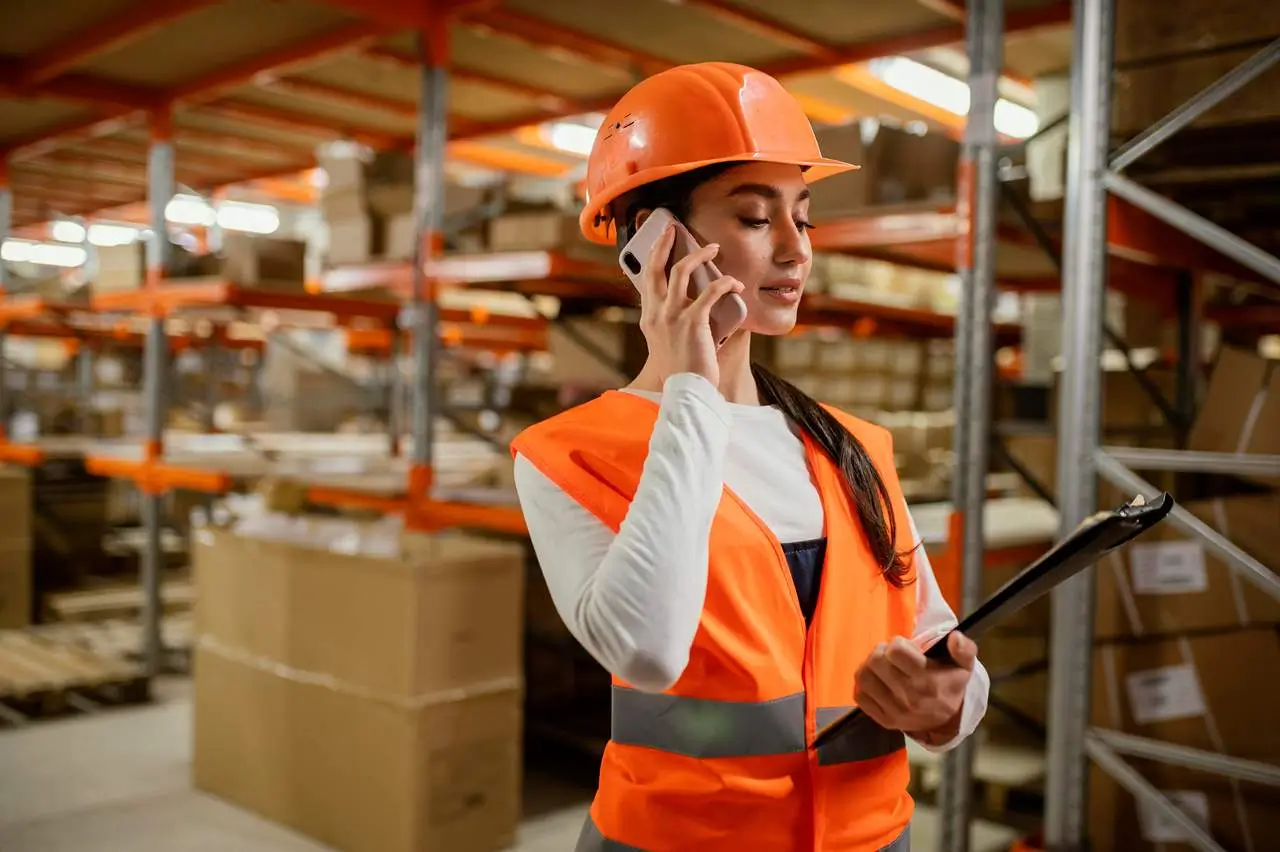 Woman in an orange helmet and vest talking on the phone while holding a clipboard in a warehouse.